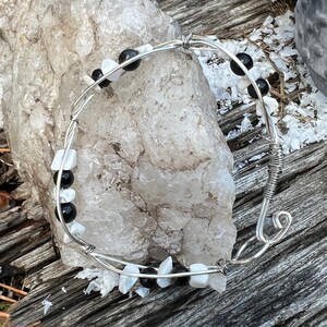 以下が含まれることがあります： 黒と白の石がついた銀色のワイヤーで巻かれたブレスレット。ブレスレットは白い岩と木製の表面の上に置かれています。