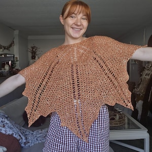 May include: A copper-coloured, hand-knitted poncho featuring an intricate, open-work lace pattern with a scalloped, leaf-like hemline. The garment is crafted from a metallic, shimmering yarn that catches the light, creating a textured, airy appearance.