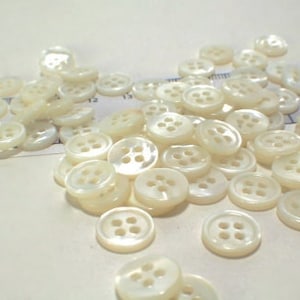 May include: A collection of small, round, white four-hole buttons scattered across a surface. A ruler is visible in the background, showing the buttons measure approximately 0.5 inches in diameter. The buttons have a smooth, pearlescent finish.