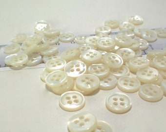 May include: A collection of small, round, white four-hole buttons scattered across a surface. A ruler is visible in the background, showing the buttons measure approximately 0.5 inches in diameter. The buttons have a smooth, pearlescent finish.