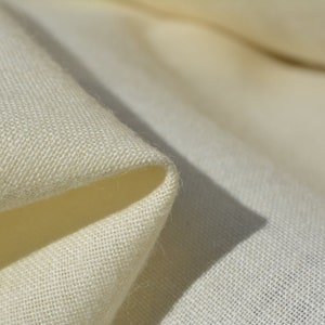 Puede incluir: Primer plano de una tela suave de color crema con una textura sutil. La tela está doblada sobre sí misma, creando una curva suave y fluida.