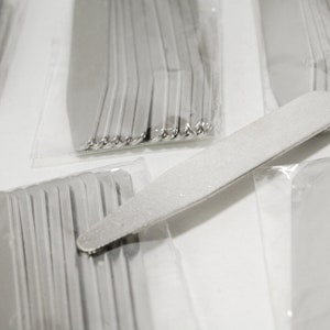 May include: A close-up of a package of white nail files. The files are arranged in a stack and are wrapped in clear plastic. The files are about 6 inches long and 1 inch wide.