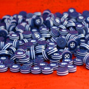 May include: A pile of navy blue buttons with white striped edges and four-hole centers. The buttons are scattered on a bright red surface, creating a visually striking contrast. These buttons are ideal for sewing and craft projects.