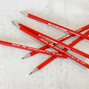 Könnte beinhalten: Eine Ansammlung roter Bleistifte mit den Marken "EDELWEISS" und "CARAN D'ACHE" auf einer hellgrauen, strukturierten Oberfläche. Die Bleistifte sind gespitzt und einsatzbereit. Der Text "SWISS MADE 341-2 HB" ist sichtbar.