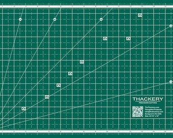 May include: A green self-healing cutting mat with white grid lines and diagonal lines. The mat has a white border with inch markings. The mat has a white label with the text "THACKERY PRECISION CUTTING" and a QR code. The label also includes the text "TheThackerry.com Thoughtful Products Designed in the USA Size: A3 13" x 19"