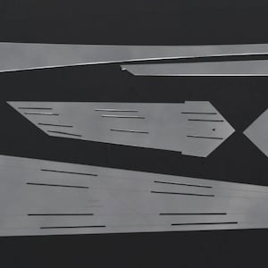 Op de afbeelding: Een verzameling zilverkleurige metalen onderdelen, mogelijk voor een knutselproject of industrieel gebruik, gerangschikt op een zwarte ondergrond. De onderdelen variëren in vorm, waaronder lange, dunne stroken en geometrische vormen met uitgesneden lijnen.