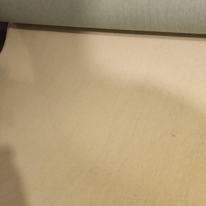 Pode incluir: Um close-up de um tecido de lona verde claro enrolado. A tela é de cor sólida com uma superfície ligeiramente texturizada. A parte inferior da imagem mostra uma tela bege plana.