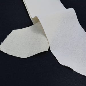 May include: Close-up of a white, textured fabric strip with a jagged edge, lying on a black surface. The fabric appears to be folded over itself, creating a layered effect. The lighting highlights the texture.