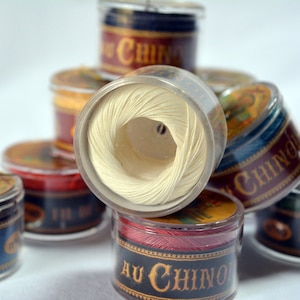 Puede incluir: Un primer plano de un carrete de hilo blanco en un recipiente de plástico transparente. El recipiente está etiquetado con "Au Chinois" en letras doradas. El carrete está rodeado de otros carretes de hilo de varios colores, incluyendo rojo, azul y verde.