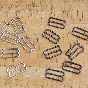 May include: A close-up of silver and black metal slider buckles. The buckles are arranged in a scattered pattern on a cork surface.