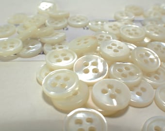 May include: A collection of off-white, round buttons with four holes each. The buttons have a pearlescent sheen and are scattered across a light-colored surface. A ruler is partially visible in the background.