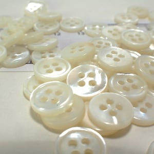 May include: A collection of off-white, round buttons with four holes each. The buttons have a pearlescent sheen and are scattered across a light-colored surface. These buttons are ideal for sewing and craft projects.