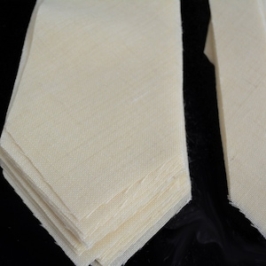 May include: A stack of off-white fabric ties. The ties are folded in half and stacked on top of each other. The ties are made of a soft, smooth fabric.