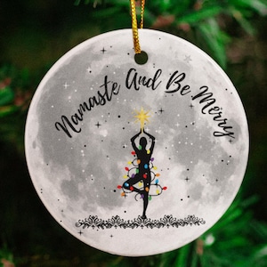 May include: A white ceramic ornament with a moon-like surface and a silhouette of a person in a yoga pose decorated with Christmas lights. The text "Namaste And Be Merry" is written around the person.