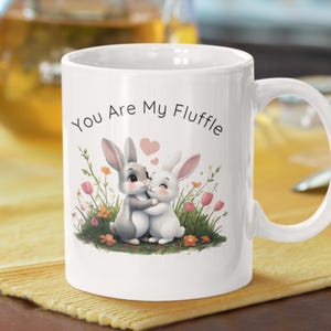May include: White ceramic mug with a curved handle. The mug features a graphic of two hugging bunnies with pink hearts and the text "You Are My Fluffle". The bunnies are surrounded by flowers and greenery.