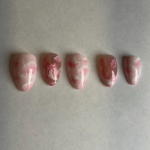 May include: Set of five almond-shaped false nails. The nails feature a pink and white cloud design, with one nail showcasing a pink bow. The nails are a light pink color with a glossy finish.
