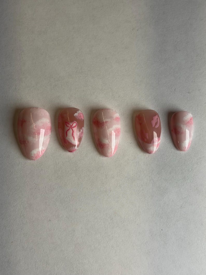 May include: Set of five almond-shaped false nails. The nails feature a pink and white cloud design, with one nail showcasing a pink bow. The nails are a light pink color with a glossy finish.