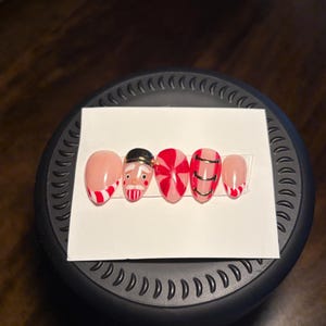 May include: A set of five artificial nails with festive Christmas designs. The nails feature a Santa Claus face, red and white candy cane stripes, and gold accents. The nails are arranged on a white card.