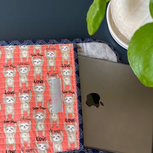 Puede incluir: Una funda para portátil de color rosa coral con un patrón repetido de perezosos colgados boca abajo. Los perezosos son marrones y tienen el texto "Lazy Days" en sus vientres. La funda tiene un borde azul marino con un detalle de costura blanca. La funda tiene un cierre de velcro. La funda contiene un portátil plateado con un logotipo de Apple negro.