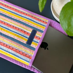 Puede incluir: Una funda para portátil de color morado con un patrón de rayas de colores. La funda tiene una solapa con cierre de velcro. Un portátil está parcialmente insertado en la funda.