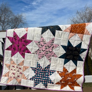 Puede incluir: Una colcha blanca con un borde morado y un diseño de patchwork de ocho estrellas. Cada estrella está hecha de una tela diferente con una variedad de patrones, incluyendo estampados florales, geométricos y de animales. La colcha está colgada en una barandilla de madera.