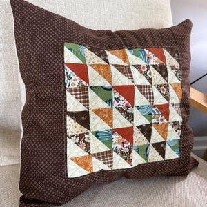 May include: Brown square pillow with a patchwork design. The center features a geometric pattern of triangles in various fabrics, including floral, plaid, and solid colors. The pillow has a dark brown background with a small cross pattern.