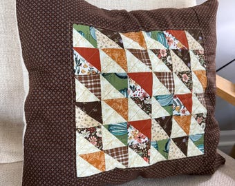 Puede incluir: Cojín cuadrado marrón con diseño de patchwork. El centro presenta un patrón geométrico de triángulos en varias telas, incluyendo estampados florales, cuadros y colores lisos. El cojín tiene un fondo marrón oscuro con un pequeño patrón de cruces.