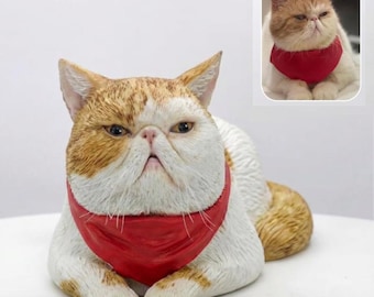 May include: A ceramic cat figurine with a grumpy expression, featuring orange and white fur and a red bandana. A smaller image of a similar cat is in the background. The cat is lying down, with its paws neatly tucked in.