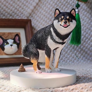 May include: A Shiba Inu dog figurine with black, white, and tan fur, posed on a white circular base. The dog has a smiling expression with its tongue out. A small brown sculpture of dog waste is next to the dog. A framed cat portrait is in the background.
