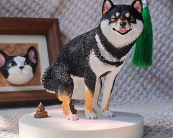 Könnte beinhalten: Figur einer Shiba Inu-Hunderasse mit schwarzem, weißem und hellbraunem Fell, die auf einer weißen, kreisförmigen Basis steht. Der Hund hat einen lächelnden Gesichtsausdruck mit herausgestreckter Zunge. Eine kleine braune Skulptur von Hundekot befindet sich neben dem Hund. Im Hintergrund ein gerahmtes Katzenporträt.