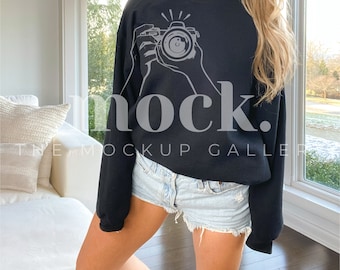 May include: A black sweatshirt with a white graphic of a camera being held by two hands. The sweatshirt is being worn by a person with long blonde hair. The person is wearing denim shorts.