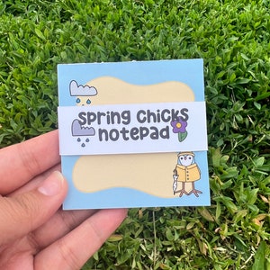 May include: A blue notepad with a yellow background and a cartoon chick wearing a yellow raincoat and holding an umbrella. The notepad has the text "spring chicks notepad" on the front.