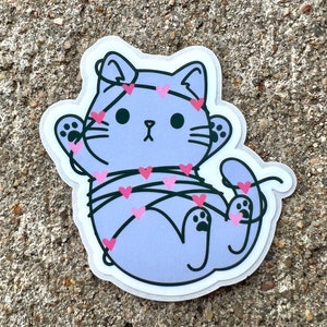 May include: A purple cat cartoon sticker with pink hearts wrapped around it. The cat has a surprised expression and is wearing a headband with hearts.