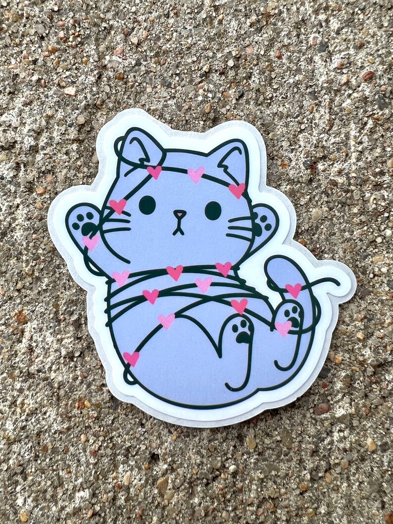 May include: A purple cat cartoon sticker with pink hearts wrapped around it. The cat has a surprised expression and is wearing a headband with hearts.