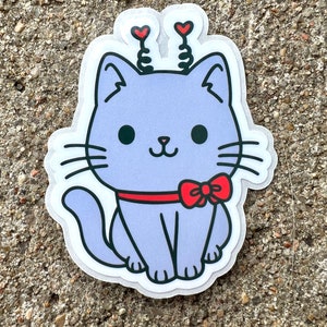 May include: A purple cat sticker with red hearts on its head and a red bow tie. The cat is sitting with its tail curled around its legs.