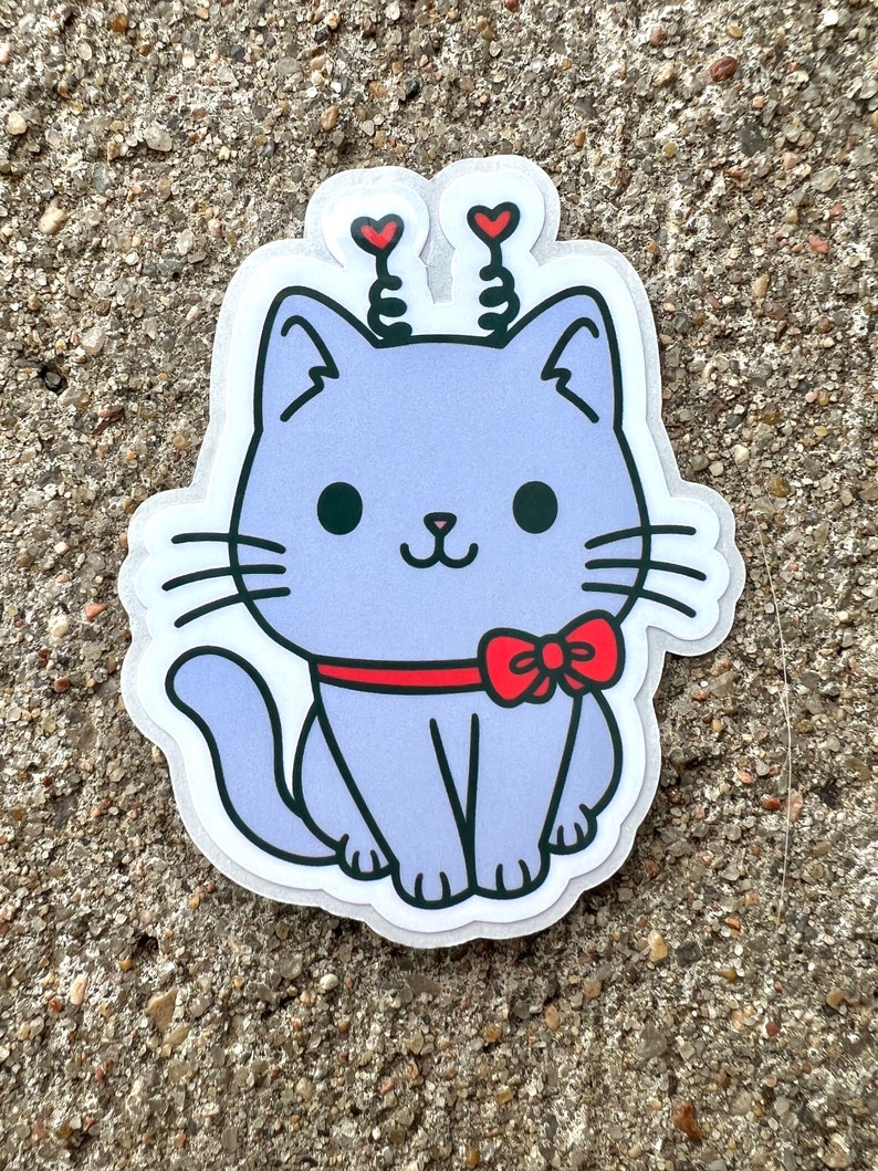 May include: A purple cat sticker with red hearts on its head and a red bow tie. The cat is sitting with its tail curled around its legs.