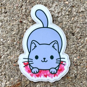 May include: A purple cat sticker with a white outline, sitting on a bed of pink hearts. The cat has black eyes and a small, pink nose.