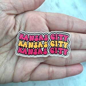 以下が含まれることがあります： レトロなバブルフォントで「KANSAS CITY」という文字が書かれた、3つの重ねられたステッカー。一番上のステッカーはピンク、真ん中のステッカーは黄色、一番下のステッカーはピンクです。