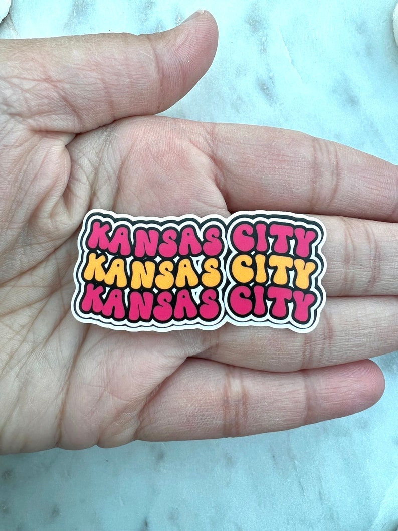 以下が含まれることがあります： レトロなバブルフォントで「KANSAS CITY」という文字が書かれた、3つの重ねられたステッカー。一番上のステッカーはピンク、真ん中のステッカーは黄色、一番下のステッカーはピンクです。