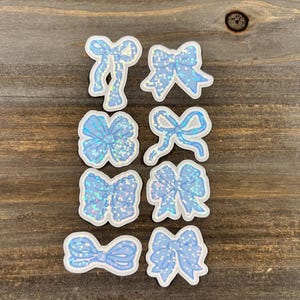 May include: A collection of eight light blue bow stickers with white outlines and glitter accents. The bows vary in shape and design, arranged on a wooden surface. These stickers are ideal for scrapbooking or crafting projects.