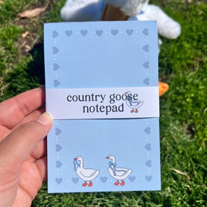 May include: A light blue notepad with a white band that reads "country goose notepad." The notepad features a border of hearts and illustrations of geese. A plush goose toy is in the background.