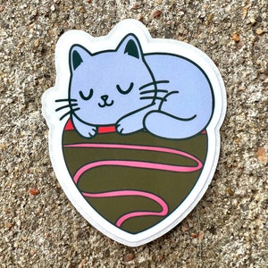 May include: A purple cat sticker with closed eyes sleeping on a brown chocolate heart with pink swirls.