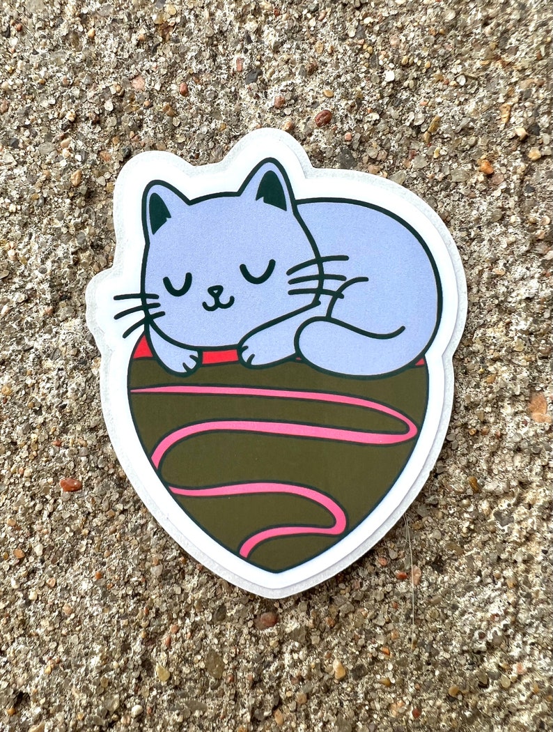 May include: A purple cat sticker with closed eyes sleeping on a brown chocolate heart with pink swirls.