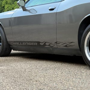 May include: A gray Dodge Challenger car with black wheels and a silver trim. The car has a black flame decal along the side with the word "CHALLENGER" in gray. The car is parked on a gray concrete surface.