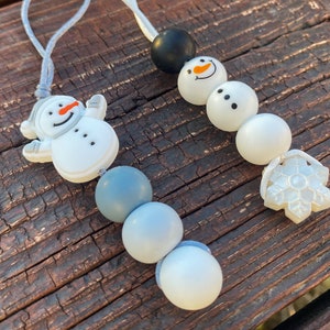 May include: Two decorative hanging ornaments featuring silicone beads. One ornament has a white snowman figure with an orange nose, followed by blue and white round beads. The other has a black bead, a snowman head, and a white snowflake charm.