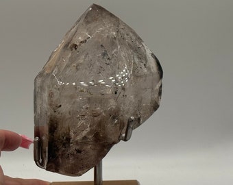 May include: A clear quartz crystal with a smoky brown hue, displayed on a wooden and acrylic stand. The crystal has a unique, faceted shape and is a beautiful example of natural beauty.
