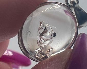 May include: A clear glass pendant with a silver bail. Inside the pendant is a small, clear crystal with a silver-colored inclusion.