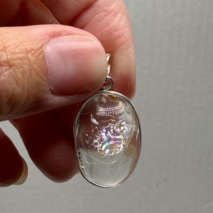 May include: An oval-shaped pendant featuring a clear, smooth, rounded stone encased in a thin silver-toned metal frame. The interior of the stone displays a reflective, iridescent patch that catches light, creating a multi-colored shimmer effect.
