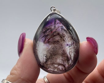 May include: A silver pendant featuring a teardrop-shaped amethyst stone with purple and grey inclusions. The stone is set in a silver bezel and has a silver bail for attaching a chain.