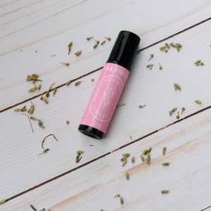 May include: A pink and black roller bottle with a label that reads "Woodland & Body Botanics" and "10 ml". The bottle is on a white wooden surface with dried lavender flowers scattered around it.